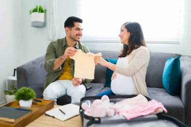 Sevimli evli bir çift bebek kıyafetlerine bakıyor ve bebeklerinin doğumu için kanepede otururken hastaneye götürmek üzere bir bavul hazırlıyorlar.
