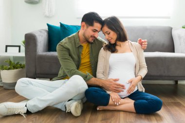 Sevgi dolu ve şefkatli bir koca hamile karısına sarılıp karnına dokunuyor. Evli bir çift, kafaları bir karış havada evde yerde oturuyorlar.