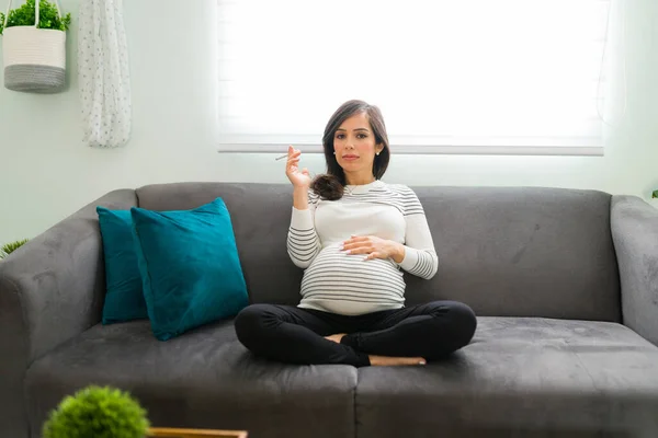 Pregnant woman smoking and holding a cigarette with a serious expression. Caucasian expectant mother with a bad and unhealthy habits for her pregnancy