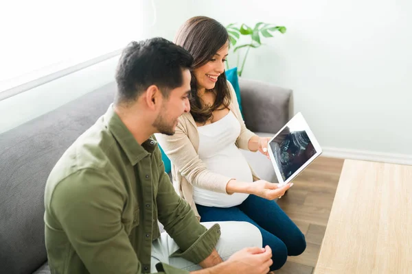 快乐的孕妇和迷人的男人笑着 一边坐在客厅沙发上一边拿着一块屏幕上有数字超声波的平板电脑 — 图库照片