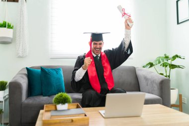 Mezuniyet elbisesiyle Latin üniversitesinden mezun olmuş. Oturma odasında otururken üniversite diploması almış. 20 'li yaşlardaki mutlu bir adam, sanal bir törenle yapılan bir video görüşmesi sırasında kutlama yapıyor.