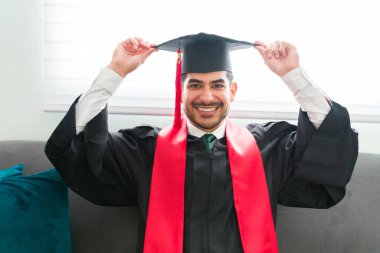 Mutlu İspanyol adam evinde otururken mezuniyet şapkası takıyor. Erkek mezun sanal seremoniye katılıp üniversite diplomasını almaya hazırlanıyor.