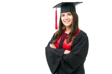 Kollarını kavuşturmuş mezuniyet elbisesi içinde poz veren gururlu genç bir kadın. 20 'li yaşlarda bir kadın üniversiteden mezun oluyor ve beyaz bir arka planın önünde duruyor.