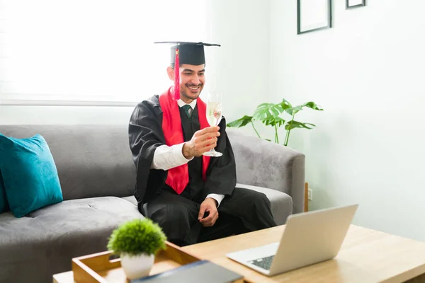 Happy latin young man making a toast with a glass of wine during his virtual graduation ceremony. Male graduate celebrating his academic success in a video call because of the coronavirus pandemic