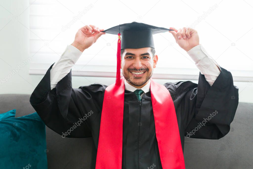 Happy hispanic man putting on a graduation cap while sitting on his home living room. Male graduate preparing to attend the virtual ceremony and receive his college diploma