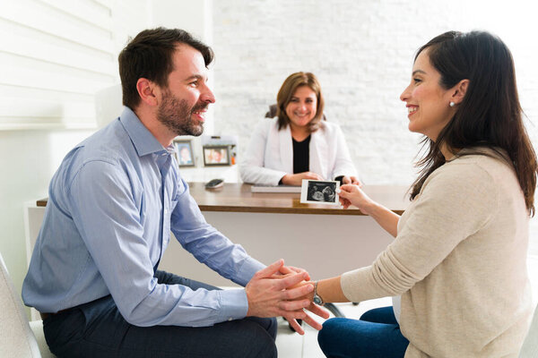 Взволнованная беременная пара, держащая отпечаток ультразвука своего ребенка, сидя за своим столом с гинекологом-женщиной. Счастливая беременная женщина и мужчина только что узнали пол своего ребенка