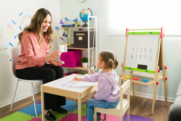 Homeschool mom teaching her little daughter how to count. Female teacher or tutor showing learning cards with numbers to an elementary girl at her home