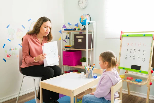 Female therapist holding a learning card with a sad face. Caucasian psychologist asking a little girl about her feelings during a therapy session