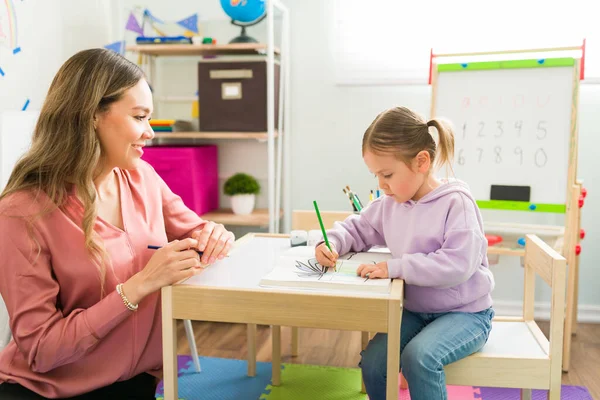 Female tutor sitting with a little elementary girl at home and looking at her. Caucasian girl learning how to draw next to her mom during a homeschool day