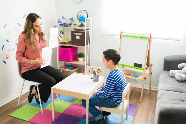 Beautiful female therapist asking a little boy about his negative or positive emotions. Psychologist showing him cards with a sad and happy face during a therapy session