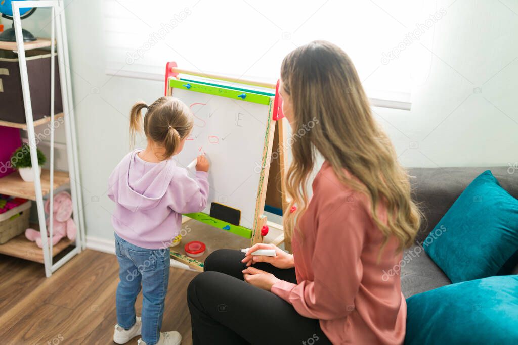 Adorable girl seen from behind writing letters on a small blackboard next to her female teacher. Caucasian tutor teaching an elementary girl the alphabet at home