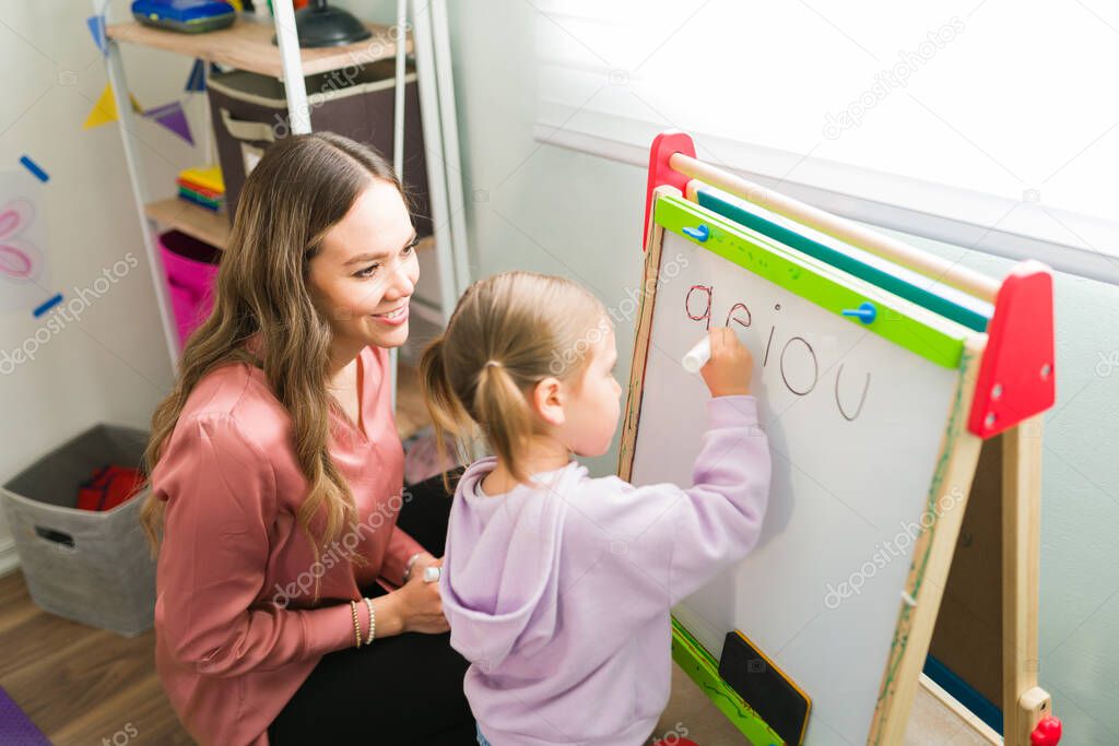 Smart elementary girl writing the vowels on a small blackboard with a female tutor. Caucasian teacher watching a little girl working during a tutoring class