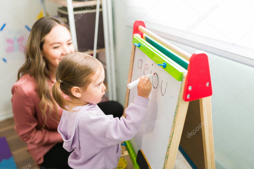 Caucasian mom smiling while watching a little girl write the alphabet on a small blackboard. Homeschool student learning how to read and write