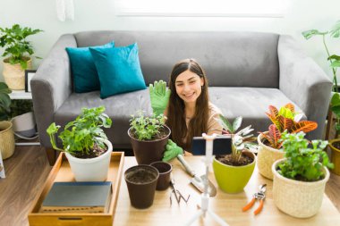 Attractive woman smiling and waving to her friends while talking on a video call with her green plants. Young woman using her smartphone to talk online about gardening 