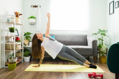 Formda genç bir kadın ev antrenmanı yapıyor. 20 yaşındaki sporcu bir kadın yan tahtada ve vücudunu yoga rutiniyle güçlendiriyor.