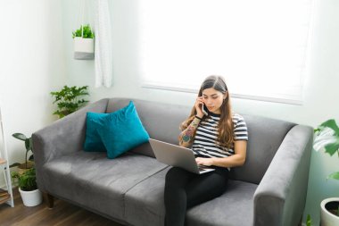 Busy hispanic young woman talking on the phone with a friend or partner while typing on her laptop. Beautiful woman working from home 