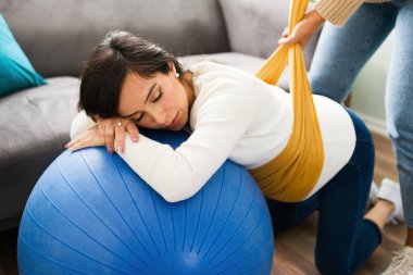 Beyaz hamile kadın nefes egzersizleri yapıyor ve doula 'sı yuvarlak karnına bir rebozo sararken bir fitness topunun üzerinde dinleniyor.