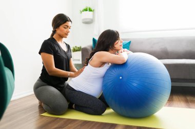 Aktif kıyafetli güzel bir kadın beyaz hamile bir kadının sırt ağrısına yardım ediyor. Müstakbel anne masaj yaptırırken spor salonunda dinleniyor.
