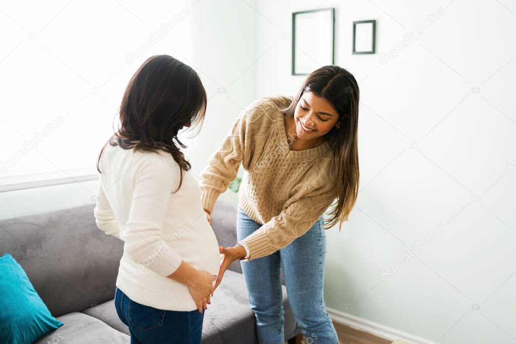 Attractive hispanic midwife smiling while touching the round belly of a caucasian pregnant woman in her home