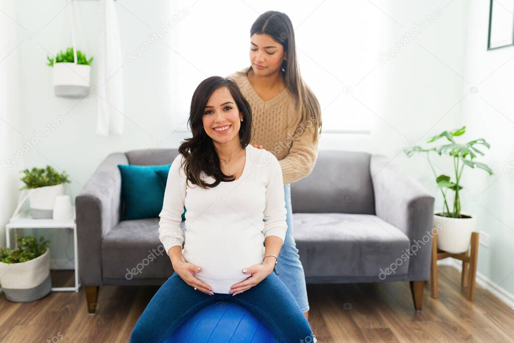Portrait of a beautiful happy pregnant woman smiling while sitting on a fitness ball and receiving a back massage from her hispanic doula