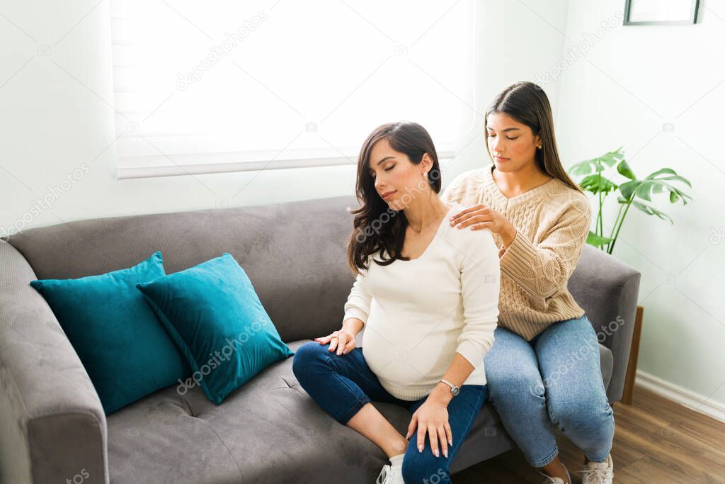 Beautiful midwife massaging the shoulders and back of a stressed pregnant woman while resting on the living room couch