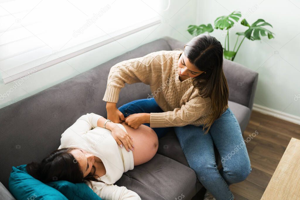 Pregnant woman and latin doula seen from above in the living room. Midwife massaging one side of the belly and hip of an expectant mother lying on the couch