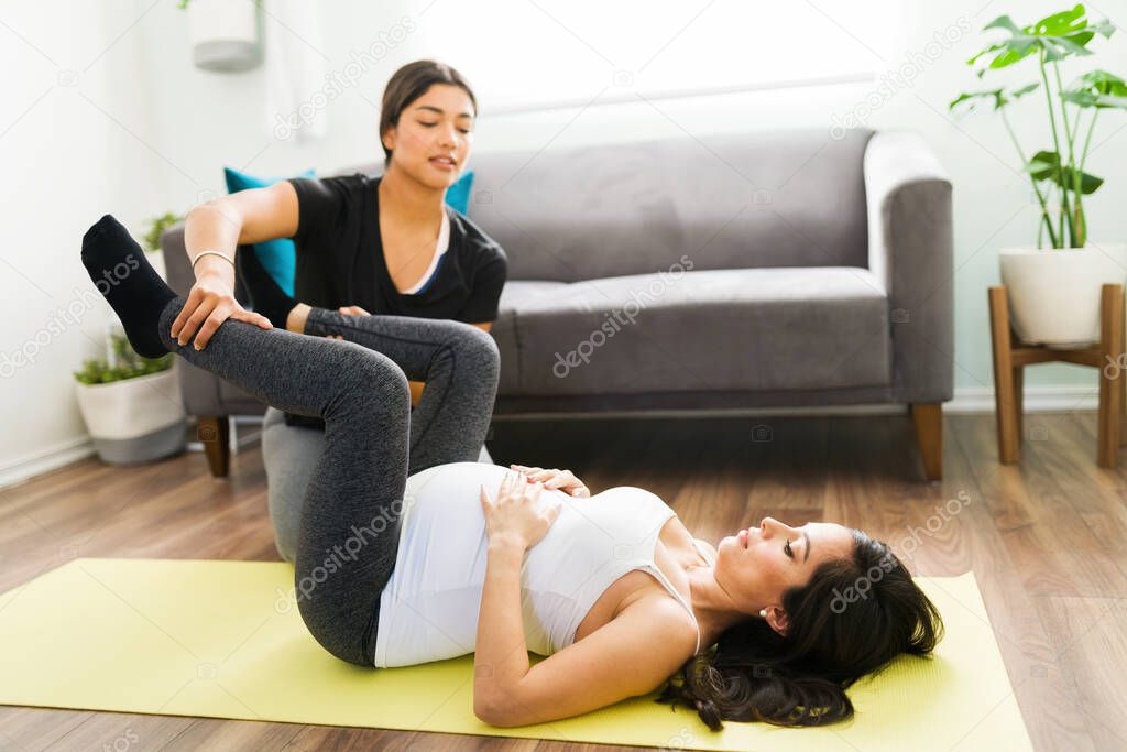 Beautiful doula teaching stretching exercises to a pregnant woman to help with her legs and hip pains. Hispanic midwife helping an expectant mother with her exercises 
