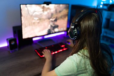Oyun bilgisayarında neon ışıklı video oyunu oynarken kulaklıklı bir kadın oyuncu arkadan görülmüş.