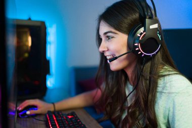 Genç bir kadın oyun bilgisayarında video oyunu oynarken çevrimiçi bir oyuncu veya arkadaşıyla kulaklık ve mikrofonla konuşuyor.