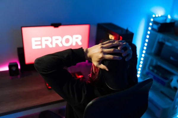 Young man seen from behind holding his head and looking surprised while watching an error message on a gaming PC after playing an online videogame