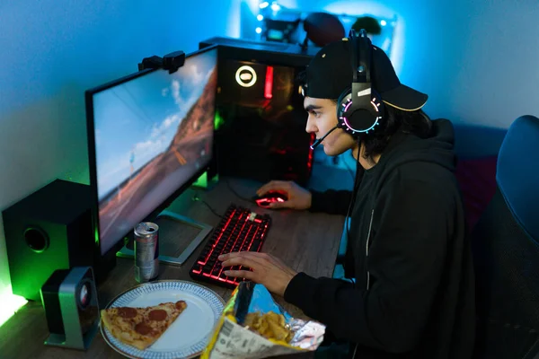 Attractive man wearing headphones and using a gaming computer with led lights to play an action video game. Young gamer eating pizza and chips while playing at his desk