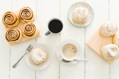 Düz tarçınlı rulo şekerler ve beyaz ahşap bir masada jöleli çörekler. Mutfak masasının üst görüntüsü hamur işleri ve kahve fincanlarıyla servis edilen tatlı.