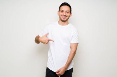 30 'lu yaşlardaki İspanyol adam gülümsüyor ve beyaz bir tişörtü işaret ediyor. Latin adam tasarım baskısı için tişört şablonu hazırlıyor