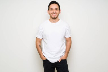 Mutlu adam elleri ceplerinde poz veriyor ve gülümsüyor. Latin adam beyaz bir arka planın önünde dikilirken tişört tasarlıyor.