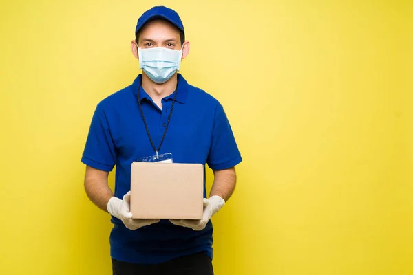 Happy man with a face mask and gloves holding a small package against a yellow background. Male courier making a delivery during the virus pandemic