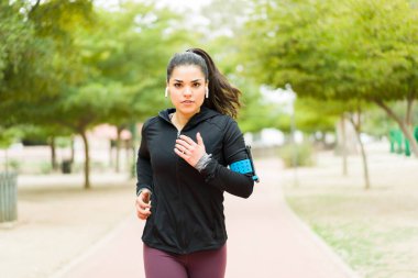 Odaklanmış ve aktif güzel bir kadın göz teması kuruyor ve kulaklık ve akıllı telefonuyla müzik dinlerken parkta koşuyor.