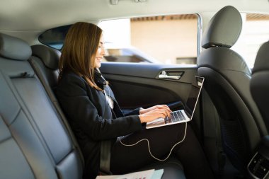 Arabasının arkasında otururken dizüstü bilgisayarında yazan neşeli bir iş kadınının yan görüntüsü. Ofise taksiyle gidip gelirken bilgisayarla gülümseyen ve çalışan beyaz bir iş kadını. 