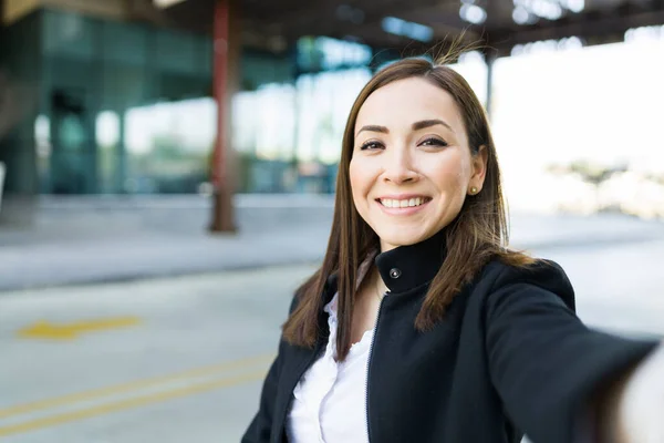 Personal perspective of a happy business woman smiling and taking a selfie with her smartphone while standing outside a modern office building