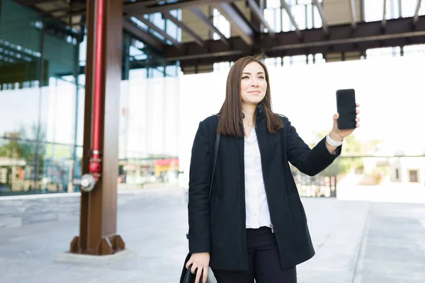 Happy business woman waiting for a taxi of a ride-share app outside her office. Professional caucasian woman smiling while holding a smartphone and a portfolio