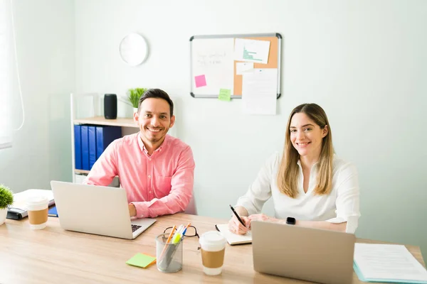 Portrait of happy professional colleagues making eye contact and smiling at the camera while working at the office with their laptops