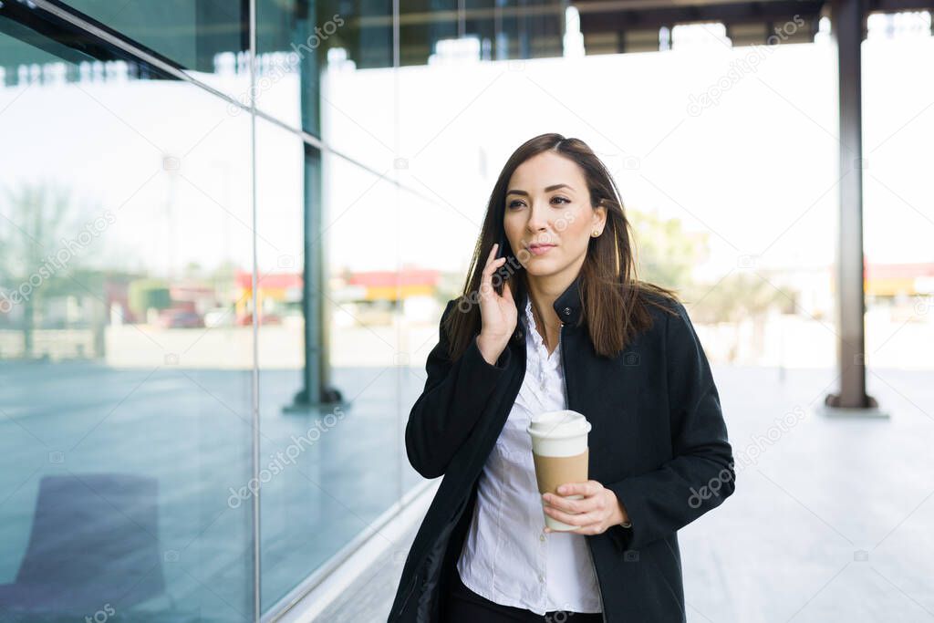 Beautiful caucasian woman in her 30s making a business call in her smartphone while walking outside a modern building and carrying a coffee