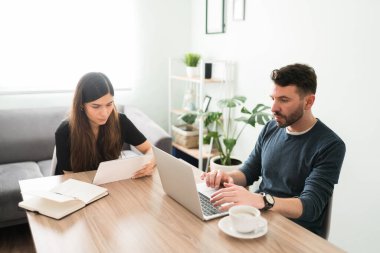 Latin kökenli genç bir çift oturma odasındaki masalarında birlikte oturuyorlar. Evde bilgisayar ve tablet kullanan çekici bir çift.