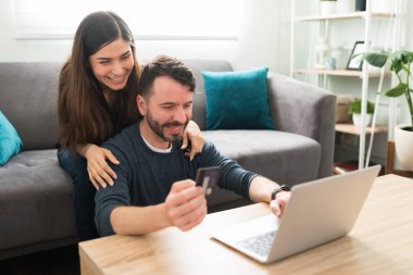 Evde dizüstü bilgisayarlarına bakarken gülen heyecanlı genç çift. Mutlu Latin sevgili ve kız arkadaş online alışverişlerini kredi kartıyla ödüyorlar.