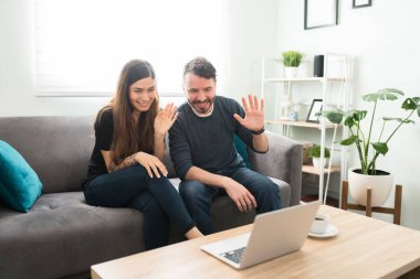 Çekici evli çift, dizüstü bilgisayarlarındaki video görüşmesi sırasında gülümseyip mutlu arkadaşlarına ve ailelerine el sallıyor.