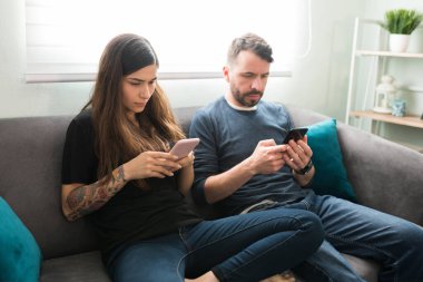 Çekici bir çift, oturma odasındaki kanepede otururken sosyal medyada akıllı telefonlarıyla mesajlaşıyor ve kaydırıyor.