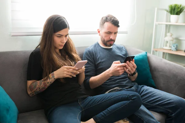 Çekici bir çift, oturma odasındaki kanepede otururken sosyal medyada akıllı telefonlarıyla mesajlaşıyor ve kaydırıyor.