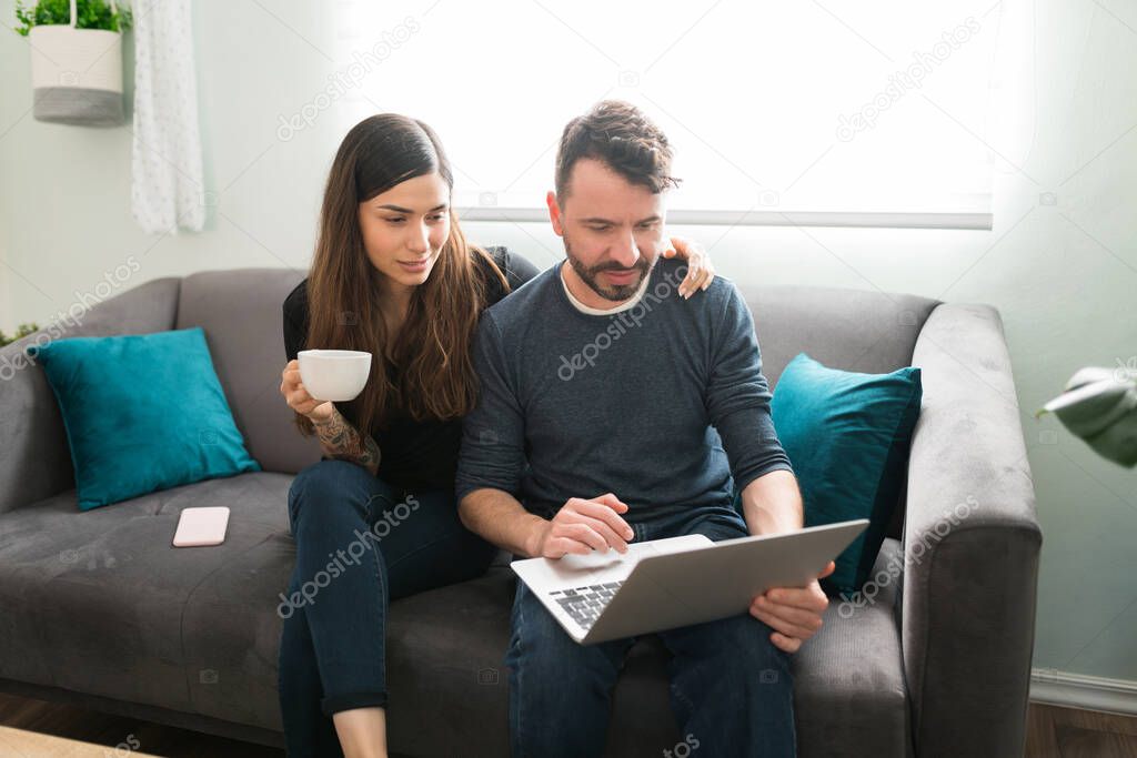 Loving wife hugging her husband while drinking coffee in the morning at home. Hispanic man showing an attractive woman his online shopping on the laptop