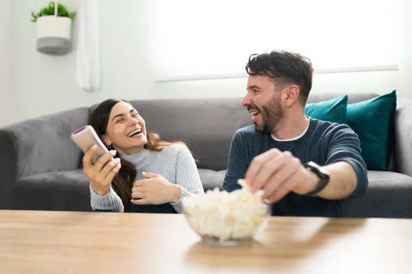 Happy couple laughing and eating popcorn while watching a comedy movie in the living room. Boyfriend and girlfriend having fun during a romantic date