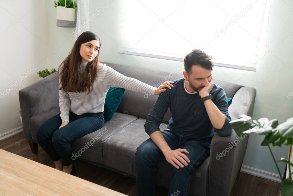 Beautiful young woman touching the shoulder of her angry handsome boyfriend while asking for forgiveness and saying sorry after having an ugly fight at home