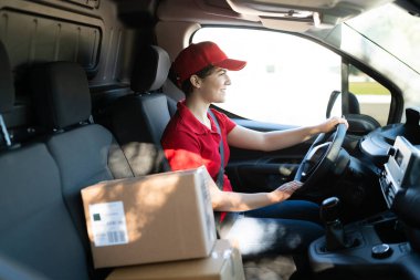 Kırmızı üniformalı ve teslimat kamyonu süren mutlu genç bir kadın. Dişi kurye, direksiyon başında otururken gülümsüyor ve paketleri teslim ediyor.